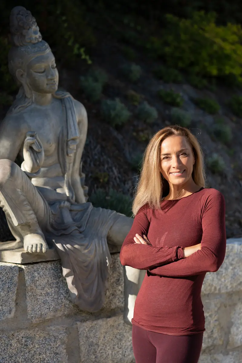 Michaela Waibel steht mit verschränkten Armen neben einer Buddha-Statue im Garten des yogamood Studios